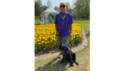 Blair and his black guide dog, Kelly, are outdoors standing on a patch of grass. Kelly is in harness and Blair is holding the harness handle.