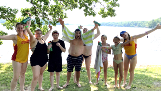 Six children and youth stand at the water’s edge wearing their bathing suits, smiling with their arms outstretched. Two lifeguards stand on opposite sides.