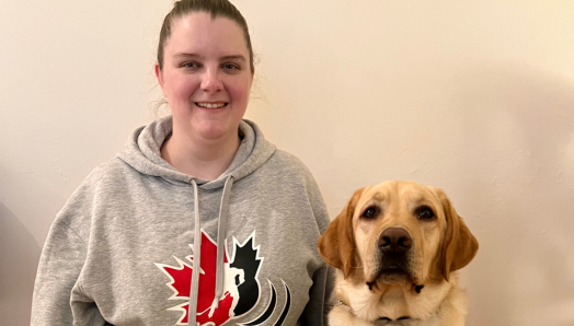 Laura and her CNIB Guide Dog, Toby. Indoors, Laura kneels on the ground next to Toby, a yellow lab in harness. Laura smiles and has her left arm around Toby. Laura is also wearing a Canadian Blind Hockey sweater. 