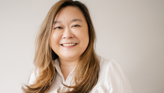 Anne Mok looking at the camera while smiling. She has long brown hair, and is wearing a white shirt.