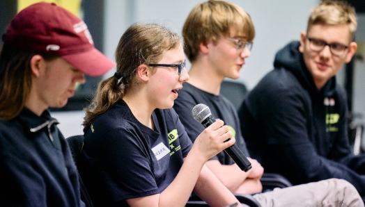 Three youths, each with light skin, sit in a row. The young girl on the left speaks into a microphone.