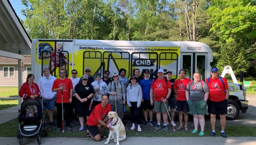 Members of the Canadian Blind Hockey Association (details) pose in front of the minibus at the CNIB Lake Joe Welcome Centre. They are wearing XX hockey jerseys and holding big hockey bags.  