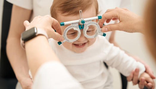 A baby is smiling while having their eyes examined. An adult is holding a measurement device up to the baby’s face.