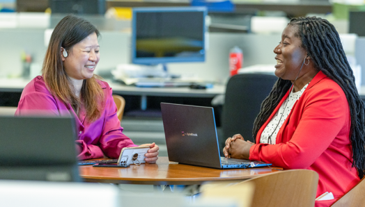 Two women sit at a table in an office environment, engaged in a conversation. Both have laptops in front of them on the table. In the background are computers and different workstations.