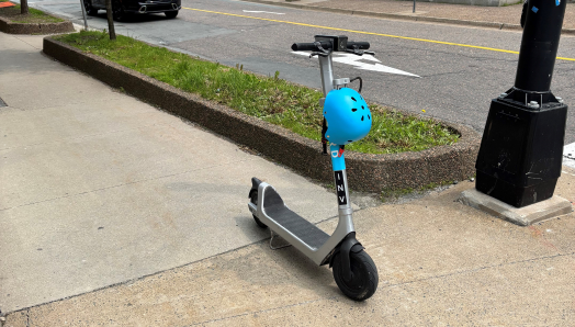A grey e-scooter parked in the middle of a pedestrian sidewalk with a blue helmet resting on the front of the scooter.