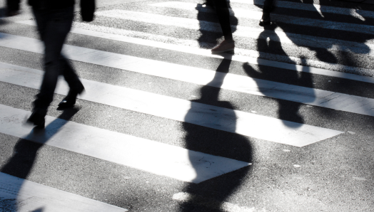 A crosswalk with shadows of people walking. The image is in black and white, showing the lower valves of several people as they cross a street. 