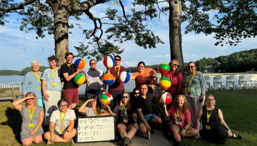 A group of CNIB Lake Joe staff and volunteers smiling outdoors on a sunny day by Lake Joseph. They are holding colourful beach balls printed with “BRL” to represent the word braille. They are posing under tall trees with white chairs and water in the background.