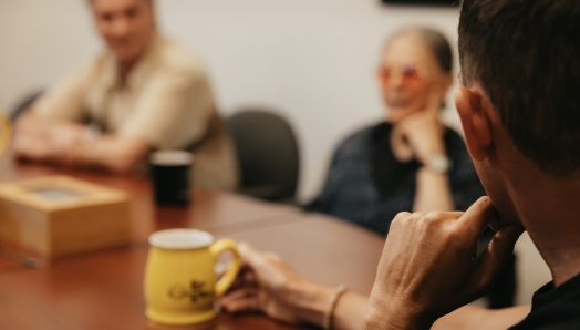 Three people sit around a conference table engaged in discussion. The focus is on a person in the foreground holding a yellow coffee mug. Two other individuals are visible in the background, slightly out of focus, one wearing red-tinted glasses and the other with arms crossed.