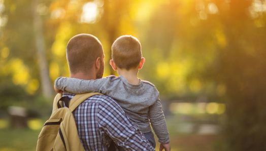 A father holds his young son. The photo is taken from behind, and the child has their arm around their father's shoulders. The background is a blurry outdoor scene with yellow and green tones.