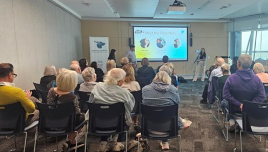 A group of people is seated in rows in a large meeting room. Shown from behind, they face two presenters at the front of the room, standing next to a screen displaying presentation slides.