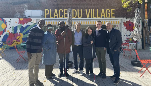  Shalom Foly, Thama Cherilus, Eitel Houédakor, Jean-Pierre Khouzam, Najla Noori, Chris McCray, and Cosimo La Rosa pose in front of Place du Village under the sun. Eitel and Najla are holding their white canes.