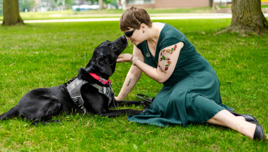 Denise and her guide dog Tara, a black Lab, sitting in the grass. Denise is gently holding Tara's face towards hers.