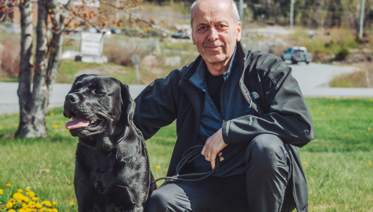 A man squats down beside a black Labrador retriever in a patch of grass. There is a tree with no leaves and a road behind them.