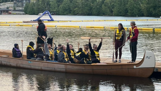 A group of people with life jackets on in a large canoe at lake joe. Everyone is smiling with their arms in the air