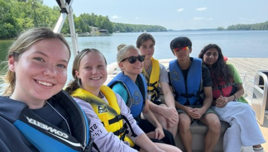 Young men and women are on a pontoon boat on the lake wearing colourful life jackets.