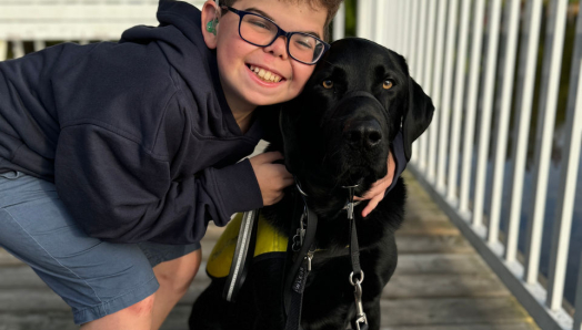 Kaiden, a young boy, is crouching down and placing his arms around a black Labrador Retriever, Flinn.