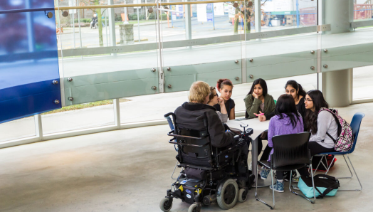 A group of seven young students sit together at a table in a classroom, participating in a shared learning activity. One of the students uses a wheelchair. 