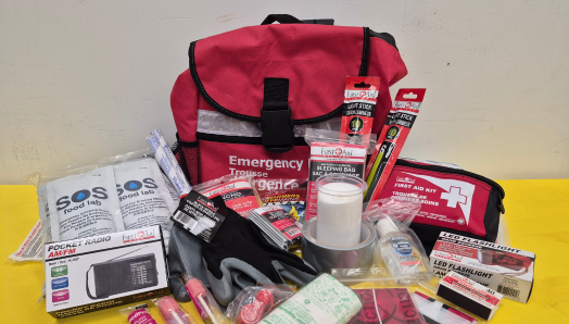 The contents of an emergency kit are shown spread out on a table. The items include an LED flashlight, emergency warming blanket, pocket radio, hand sanitizer, SOS food packets, and more.