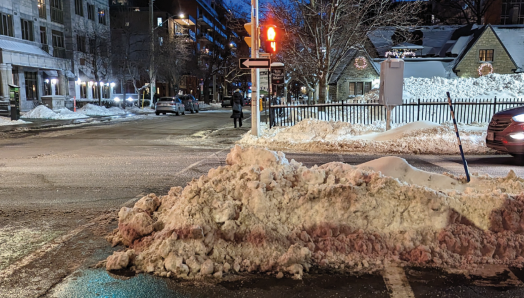 A pedestrian crosswalk in an urban neighbourhood is shown on a winter evening. The crosswalk is blocked by a snowbank piled several feet high.