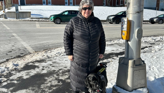 Marisa and her guide dog, a black lab named Harley, are out on a sunny winter day standing at a crosswalk next to a traffic utility pole that includes a new APS system.