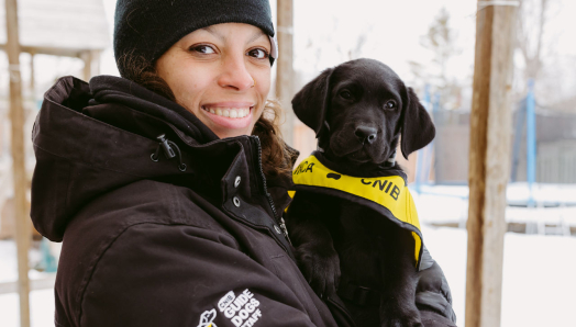 A smiling person in a winter jacket holds a black puppy wearing a CNIB Future Guide Dog vest.