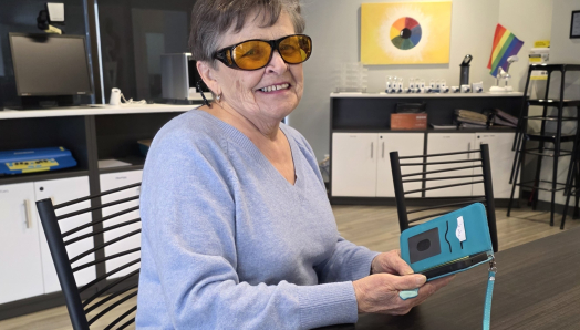 An older woman wearing amber-tinted glasses holding an accessible smartphone in a teal case.