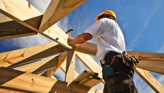 Construction worker wearing a hard hat uses a hammer to secure wooden roof beams, viewed from below against a bright blue sky.