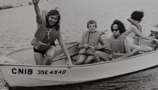 A black and white photo of two women and two children wearing lifejackets. They are smiling and sitting in a boat with CNIB on the side in the lake