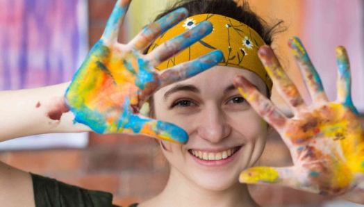 A young woman is smiling at the camera. Her hair is pulled back by a gold flowered bandana. She is holding her hands open in front of her, and they are covered in bright paint colours.