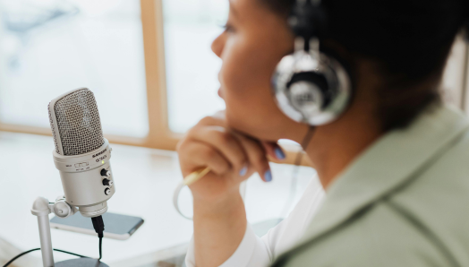 A woman wearing over-ear headphones sits at a desk, positioned beside a microphone on a stand. They are recording a podcast. 