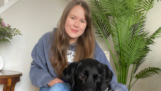 Taylor, a young woman with long brown hair, sits in a chair with her arm around her guide dog Wallace, a black lab wearing a CNIB harness, who sits beside her.