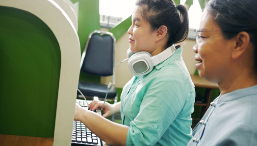 In an office cubicle, a woman sits at a computer workstation and types on a keyboard. A pair of large headphones rests around her neck. Next to her, a colleague sits at another workstation. They are reviewing something work-related on the computer monitor.