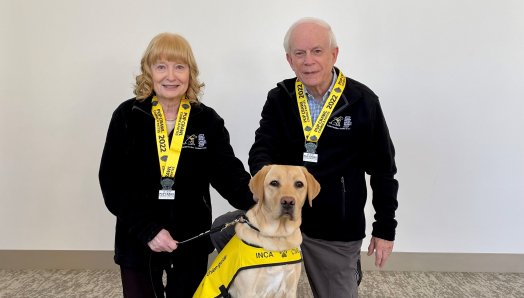 Mary and John Crocker, both smiling, kneel on the ground next to a blonde dog wearing a CNIB "Future Guide Dog" vest