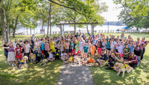 A large group of people of all ages, including many who are accompanied by buddy dogs or holding white canes, raise their hands in the air joyfully at Lake Joe - with the lake visible behind them.