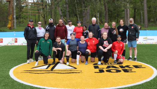 A group of blind soccer players of varying heights, ages, and skin tones stand in athletic gear on the accessible soccer pitch at CNIB Lake Joe, with a blind soccer ball in front of them.