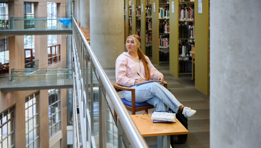 Ashlyn, wearing a pink sweater and jeans, sits in a wooden chair beside the glass railing of a multi floored library. She has a notebook and pen on her lap and is looking out over the railing's edge.