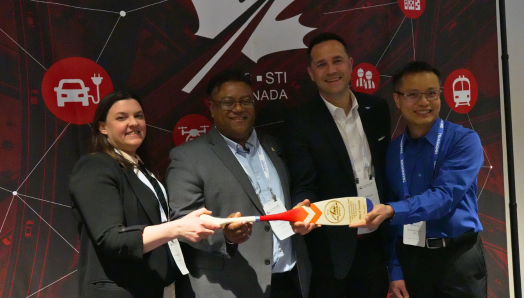Four members of the Accessible Sidewalk Construction Zones for Blind and Low Vision Pedestrians pilot project team stand together at an event, smiling and holding their award, a wooden paddle engraved with the award information. Mary Ann, far left, holds one end of the paddle while the other team members gather around it.
