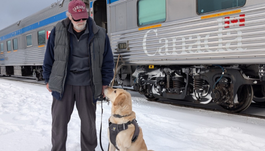 Dan stands in the snow beside his yellow Labrador guide dog, Sparky, who sits in harness and looks up at him. A VIA Rail passenger train is stopped on the tracks behind them.