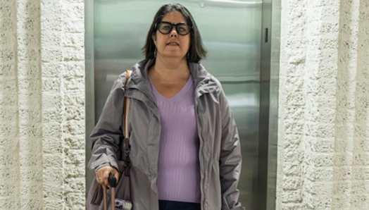 Karoline stands next to her black Labrador guide dog, Guardian, in front of an elevator. Guardian is in harness; Karoline holds the harness handle in her left hand and the handle of a suitcase in her right hand.