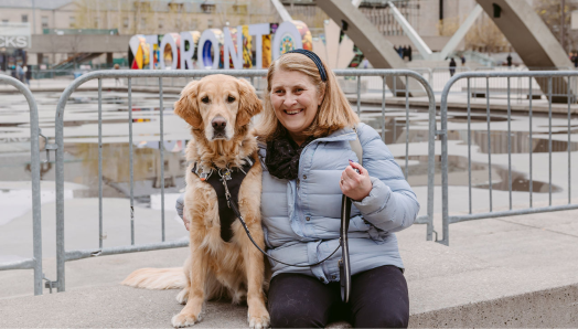 Lindsay sits beside her golden retriever guide dog, Charles, at Nathan Phillips Square in Toronto. Charles is in harness, and Lindsay smiles at the camera with one arm around him. 