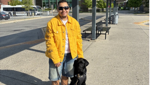 Tye stands beside his black Labrador guide dog, Zed, at an outdoor transit station. Tye hold’s Zed’s harness and leash. 