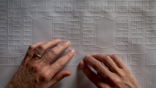 Two hands move across a large tactile architectural floor plan. Raised lines outline rooms, corridors, walls, and other features of the building layout.