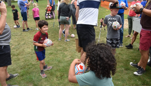 A crowd of children and adults gather on a grassy lawn for a soccer activity. Many of them are holding small white soccer balls in their hands or kicking them around their feet.