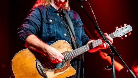 Alan Doyle smiles at the audience while performing onstage with an acoustic guitar at a microphone.